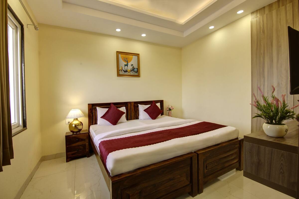 Quarto de hotel com cama de madeira, mesa de cabeceira com abajur, um quadro na parede e planta em vaso sobre um aparador de madeira.