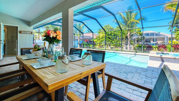 A dining area with a view of a swimming pool, a sign reading "Beach House", and a table set with a vase of flowers.