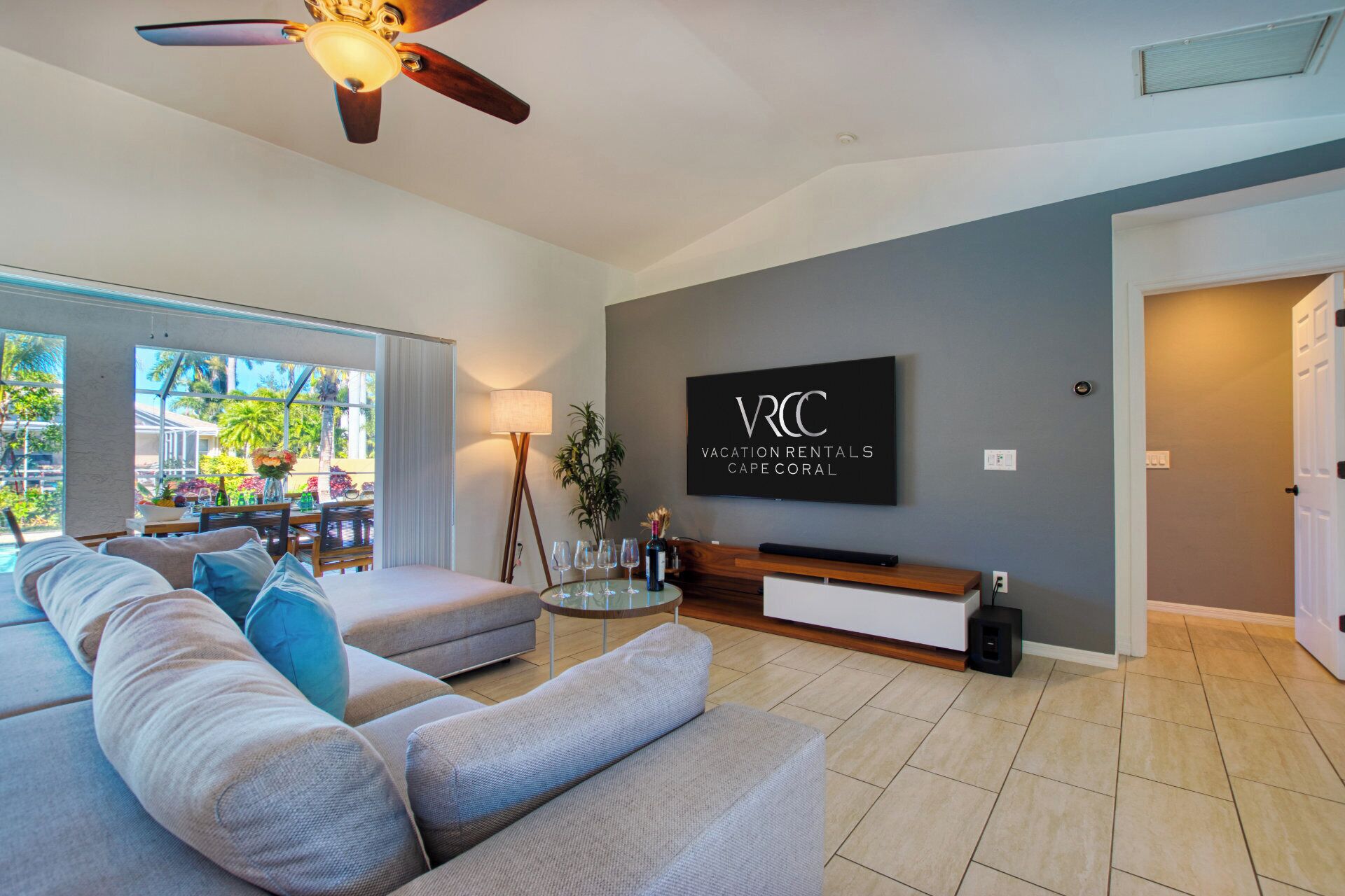 Un salon moderne avec un canapé gris, un ventilateur de plafond et un téléviseur affichant le message « LOCATION DE LIEUX POUR LA VACCINATION À CAPE CORAL ».