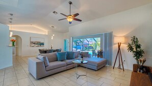 A modern living room with a sectional sofa, a coffee table, and a ceiling fan. There is a dining area with a table and chairs, and a view of a pool through large windows.