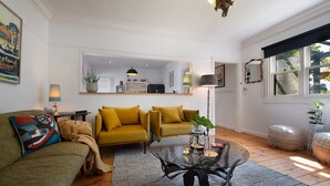 A living room with a sofa, a coffee table, and a lamp. There is a window with a view of trees and a door leading to another room.
