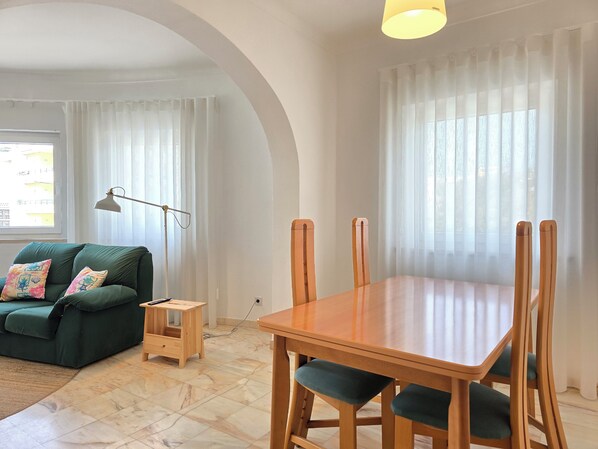 A living room with a green sofa, a wooden coffee table, and a dining table with wooden chairs.