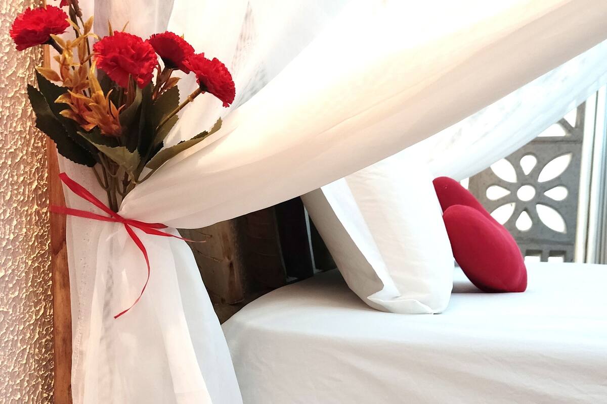 A bed with white bedding and a red pillow, a bouquet of red flowers tied with a red ribbon, and sheer white curtains.
