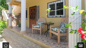 Een overdekt terras met twee houten stoelen en een kleine tafel, een houten deur en een raam met decoratieve ijzeren tralies.