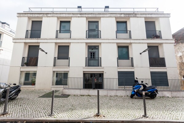 A multi-story building with balconies, a gated entrance, and a paved ground.