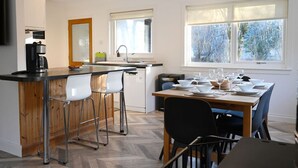 A modern kitchen with a central island, bar stools, a dining table set for six, and large windows with a view of trees. - Rivendell House located close to Fort William (Spean Bridge)