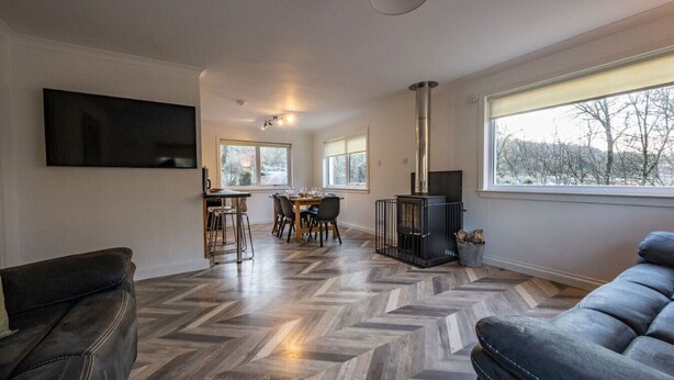 A modern living room with a herringbone floor, a fireplace, a dining area with a table and chairs, and a large window providing a view of the outdoors. - Rivendell House located close to Fort William (Spean Bridge)