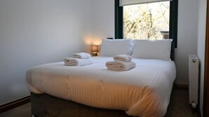 A neatly made bed with towels, a bedside lamp, and a window with blinds. - Rivendell House located close to Fort William (Spean Bridge)