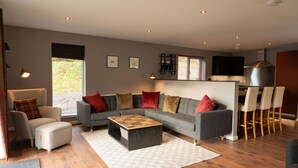 A modern living room with a grey sofa, red and beige cushions, a wooden coffee table, and a white armchair. There is a kitchen area with bar stools and a large stove. - Cairn Cottage in the village of Onich (Onich)