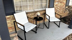 Two white chairs with black metal frames and a small round table with a vase of flowers, positioned on a concrete patio in front of a brick building with large windows and shutters. - Agreeable Gray: 3 Bedroom, Soothing 5 Beds, Large groups, Pet Friendly

 (Tyler)