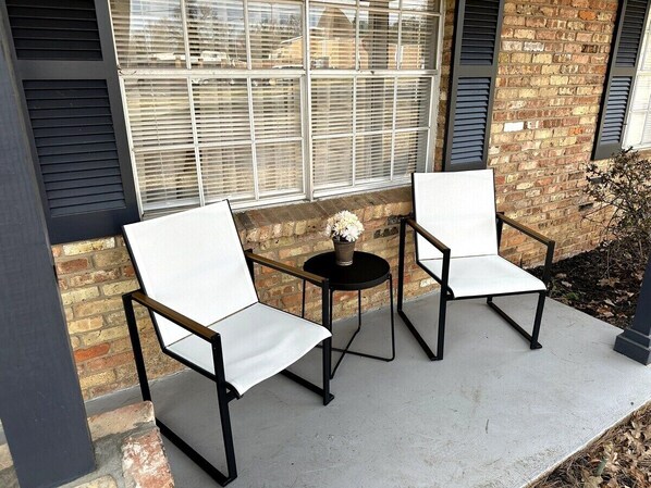 Two white chairs with black metal frames and a small round table with a vase of flowers, positioned on a concrete patio in front of a brick building with large windows and shutters. - Elegant 3 BR House, Fenced Yard, Crews, Couple, Relaxing Getaway (Tyler)
