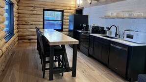 A log cabin interior with a wooden dining table, chairs, a kitchen with modern appliances, and a window with a view of a car outside.