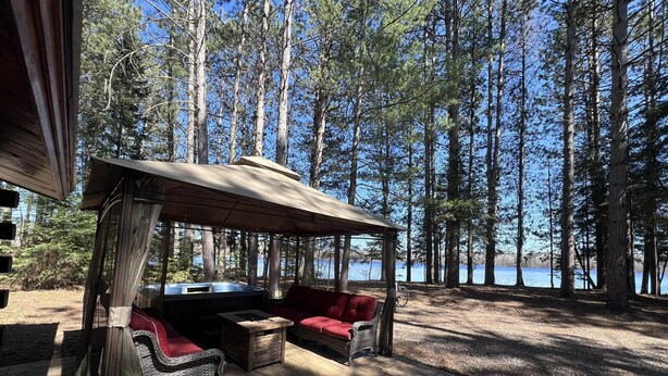 A covered outdoor seating area with wicker furniture and a fire pit, overlooking a forest and a lake.