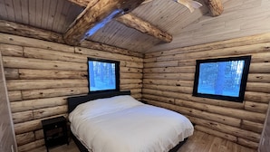 A log cabin bedroom with a bed, two windows, and a ceiling fan.