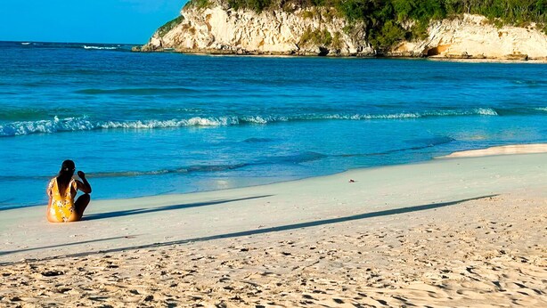 A beach with clear blue water, a sandy shore, and a rocky cliff with greenery.