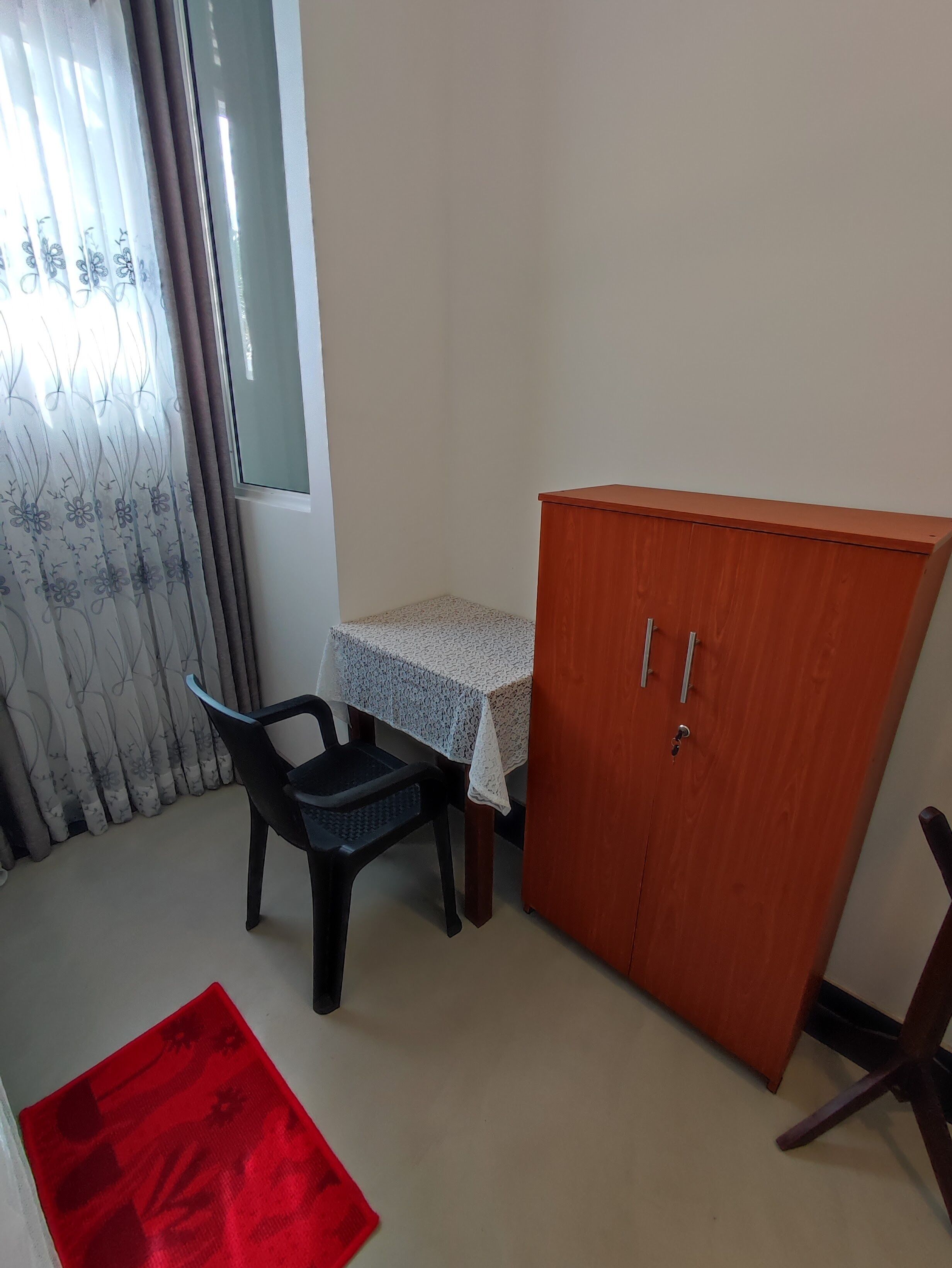 A small room with a wooden cabinet, a table with a lace tablecloth, a black chair, and a red rug.