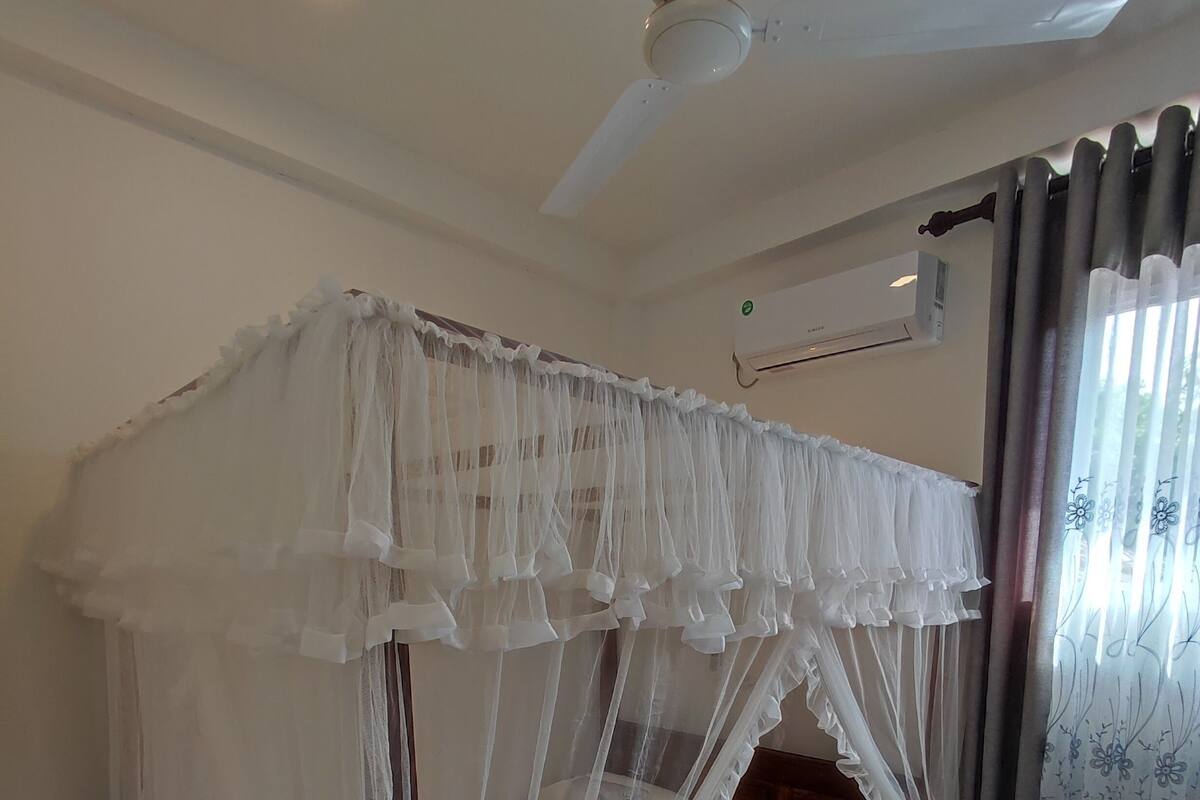 Un dormitorio con cama con dosel, ventilador de techo, aire acondicionado, alfombra roja y una silla.