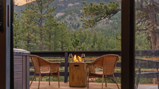 A wooden deck with wicker chairs and a wood-burning stove, overlooking a mountainous landscape. - Elegant A-Frame w/ Hot Tub Views! Close to town, skiing, & hiking! (Evergreen)