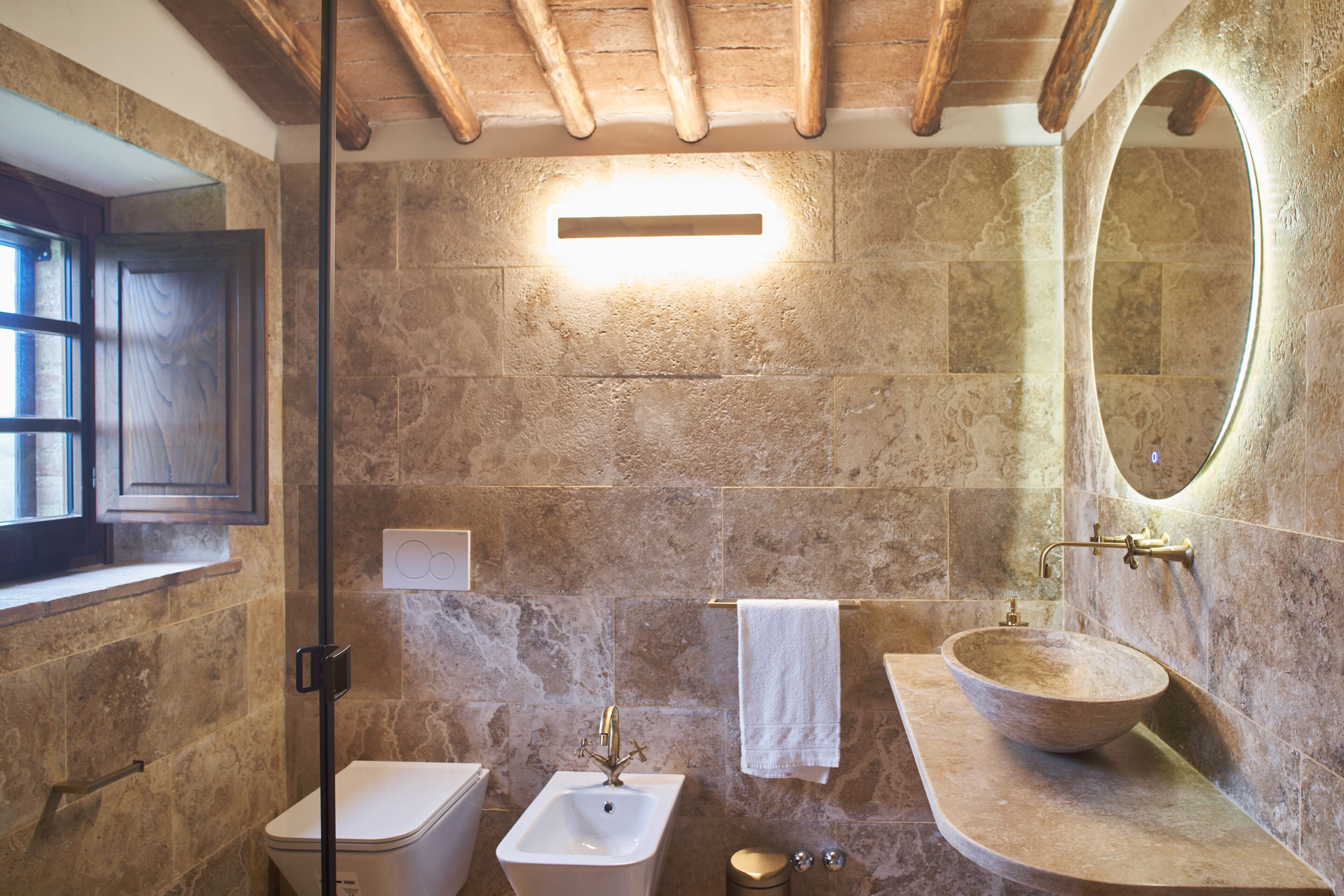 A bathroom with a stone wall, a wooden ceiling, a large oval mirror, a wooden basin, and a white towel hanging on the wall.