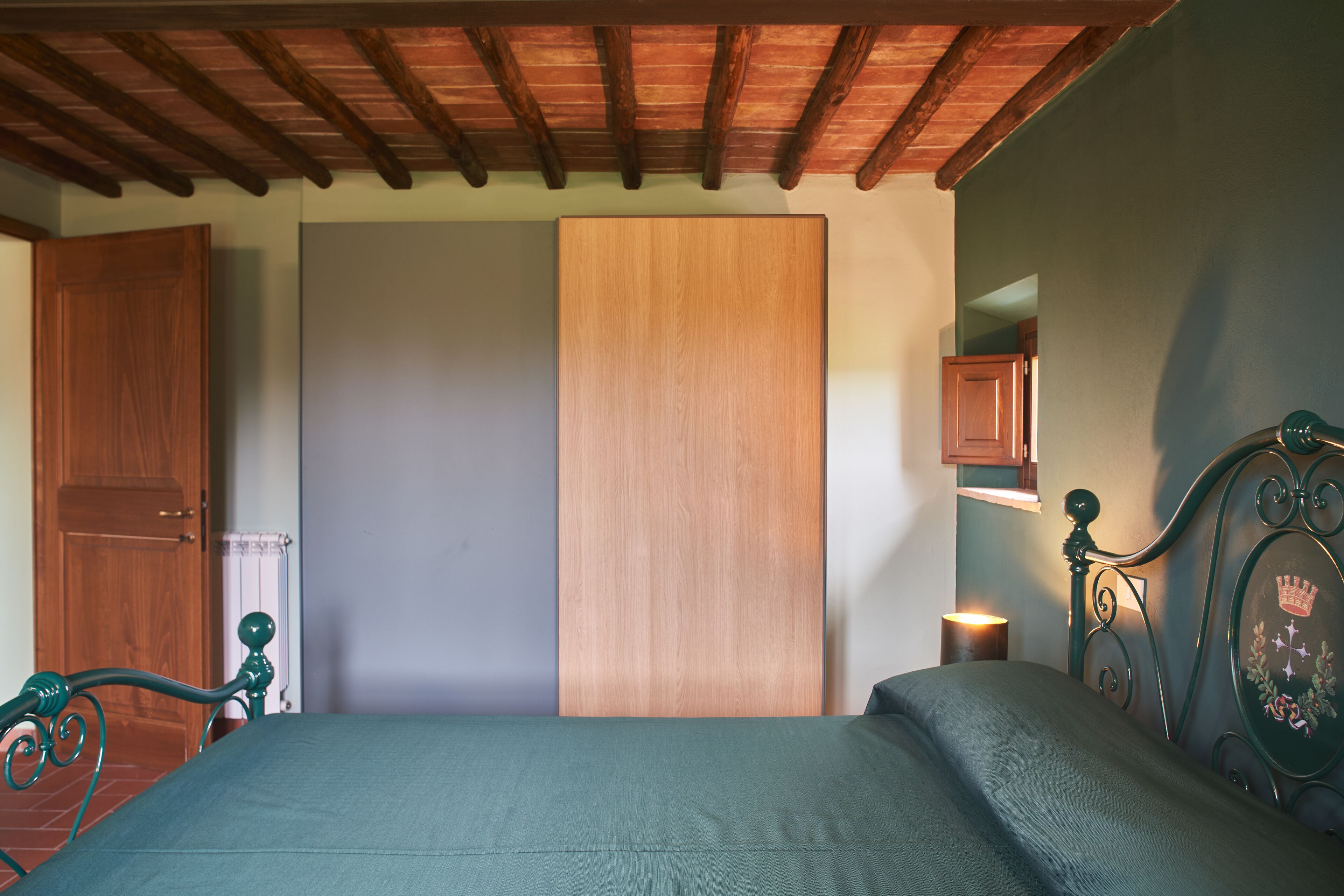 Une chambre avec un plafond en bois, un lit recouvert d’une couverture verte, une table de chevet avec une lampe et une armoire en bois.