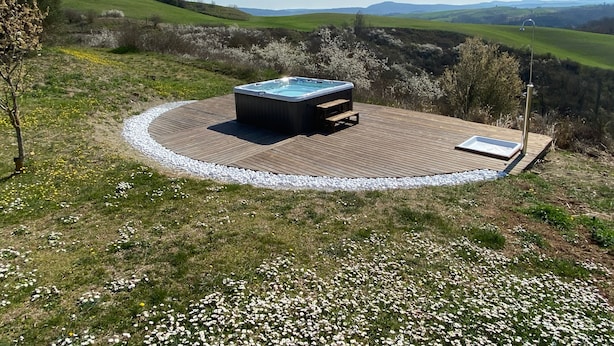 A hot tub on a wooden deck surrounded by a white gravel border, set in a grassy area with a view of rolling hills in the background. - Podere Sant'Antonio (Asciano)