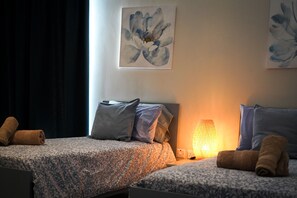 Une chambre d’hôtel avec deux lits, chacun recouvert d’une couvre-lit à motifs, agrémenté d’oreillers et de serviettes enroulées. Une lampe est fixée au mur et un tableau est accroché au-dessus des lits.