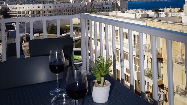 Un balcón con dos copas de vino y una planta en maceta, con vistas a una ciudad costera.