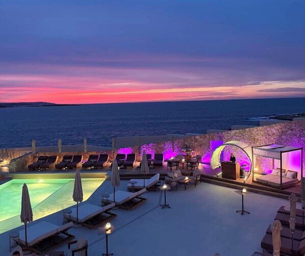A rooftop pool area with lounge chairs and a bar, overlooking the sea.