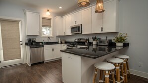 A modern kitchen with white cabinets, a central island with stools, stainless steel appliances, and a window with blinds.