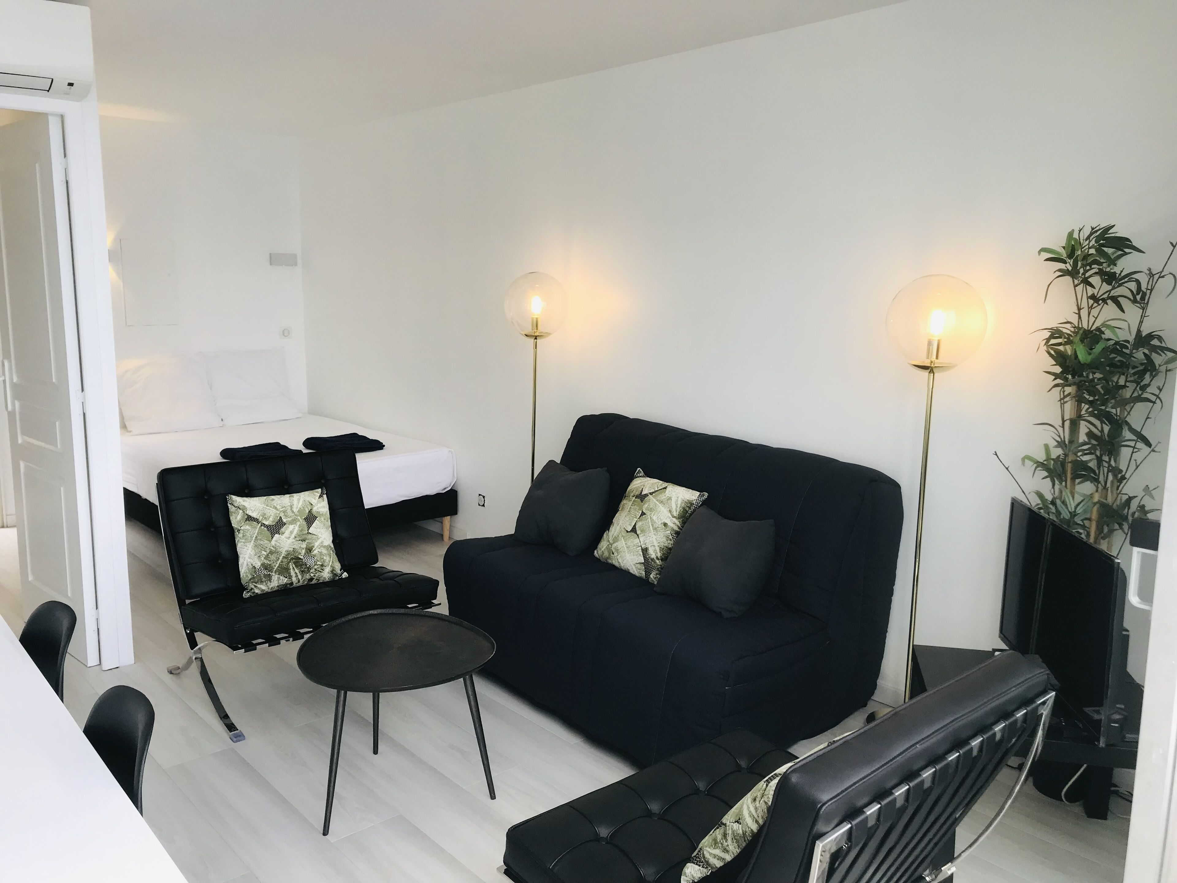 Una sala moderna con un sofá negro, dos sillones negros, una pequeña mesa redonda y una cama con ropa de cama blanca. Dos lámparas de pie proporcionan luz.