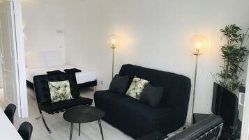 Una sala moderna con un sofá negro, dos sillones negros, una pequeña mesa redonda y una cama con ropa de cama blanca. Dos lámparas de pie proporcionan luz.
