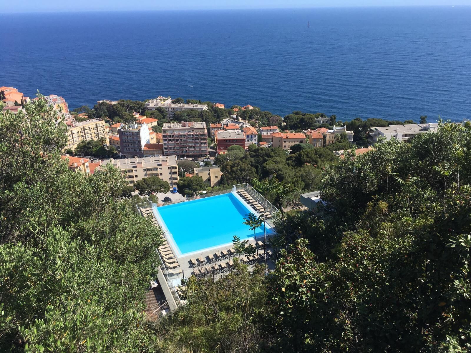 A hotel with a large swimming pool surrounded by lush greenery, overlooking a coastal area with buildings and the sea.