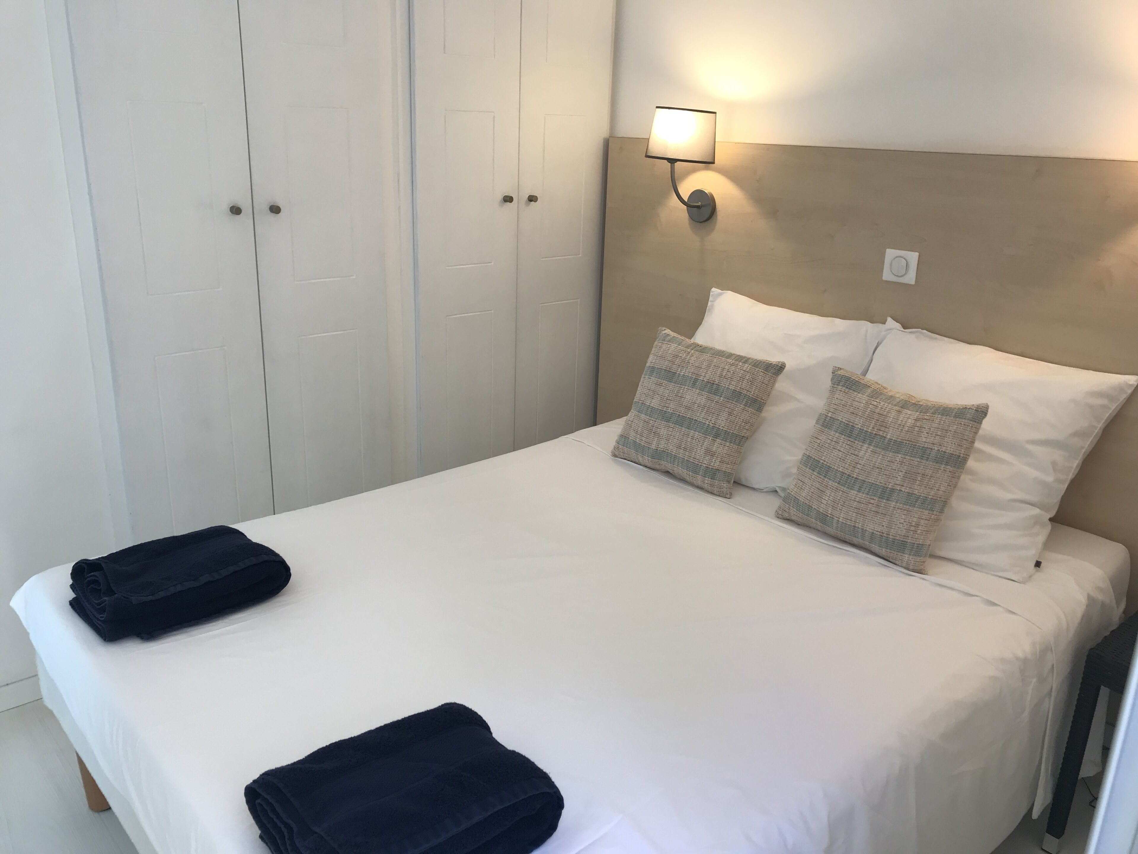 A neatly made bed with two pillows, a bedside lamp, and folded towels on a white bedspread.