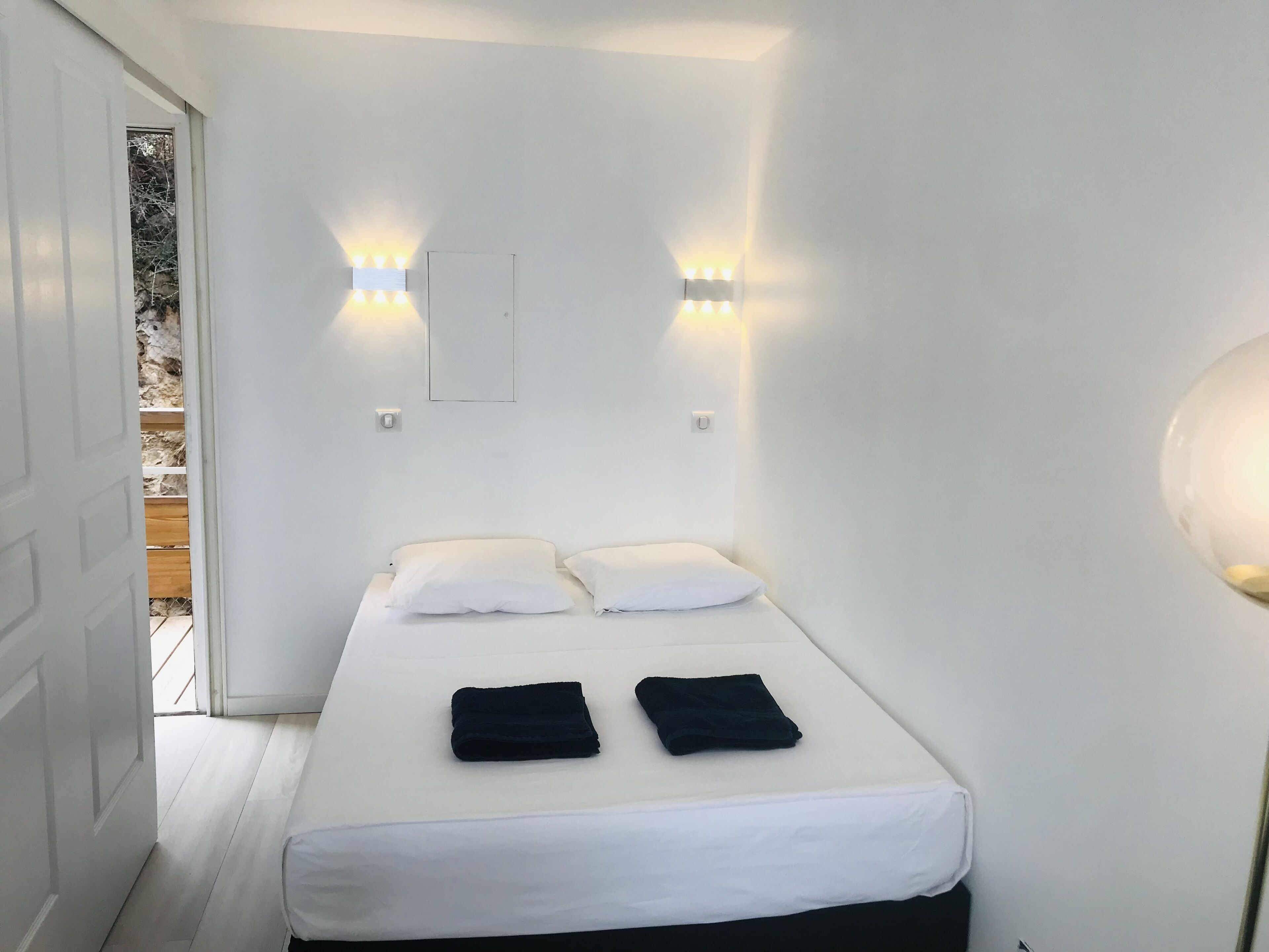 Un lit simple avec une literie blanche et deux serviettes noires, une porte blanche et un luminaire fixé au mur.