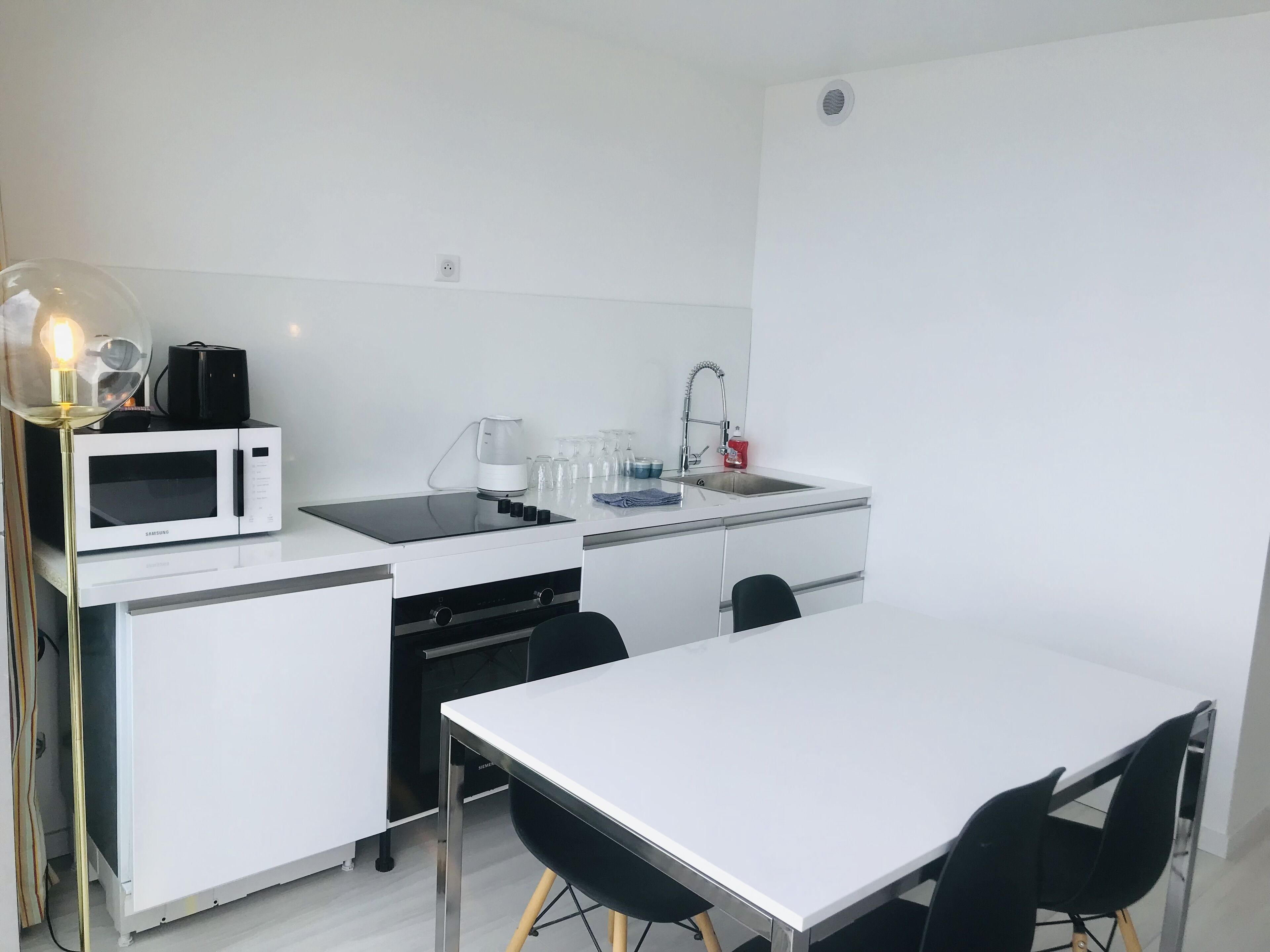 Une petite cuisine équipée d’un micro-ondes, d’une bouilloire et d’un évier. On y trouve également une table avec des chaises et une lampe.