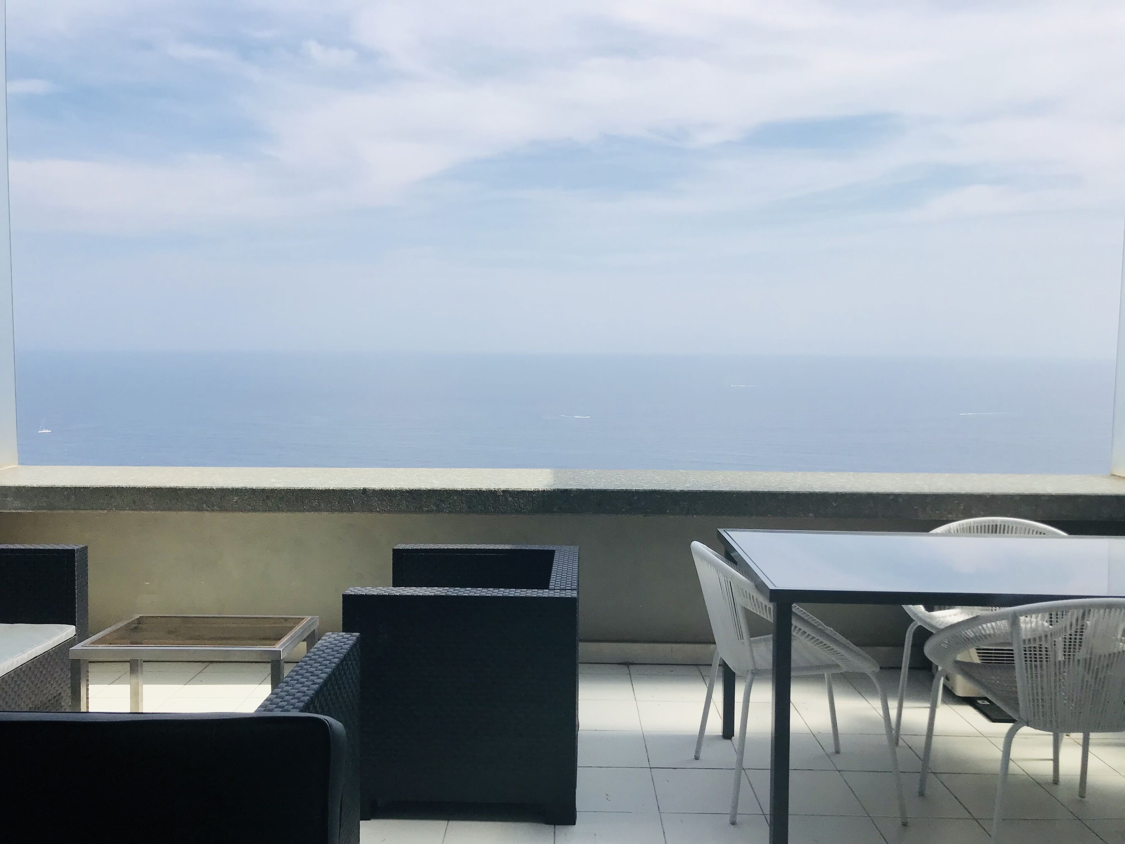 A terrace with a view of the sea, featuring black and white wicker furniture and a tiled floor.