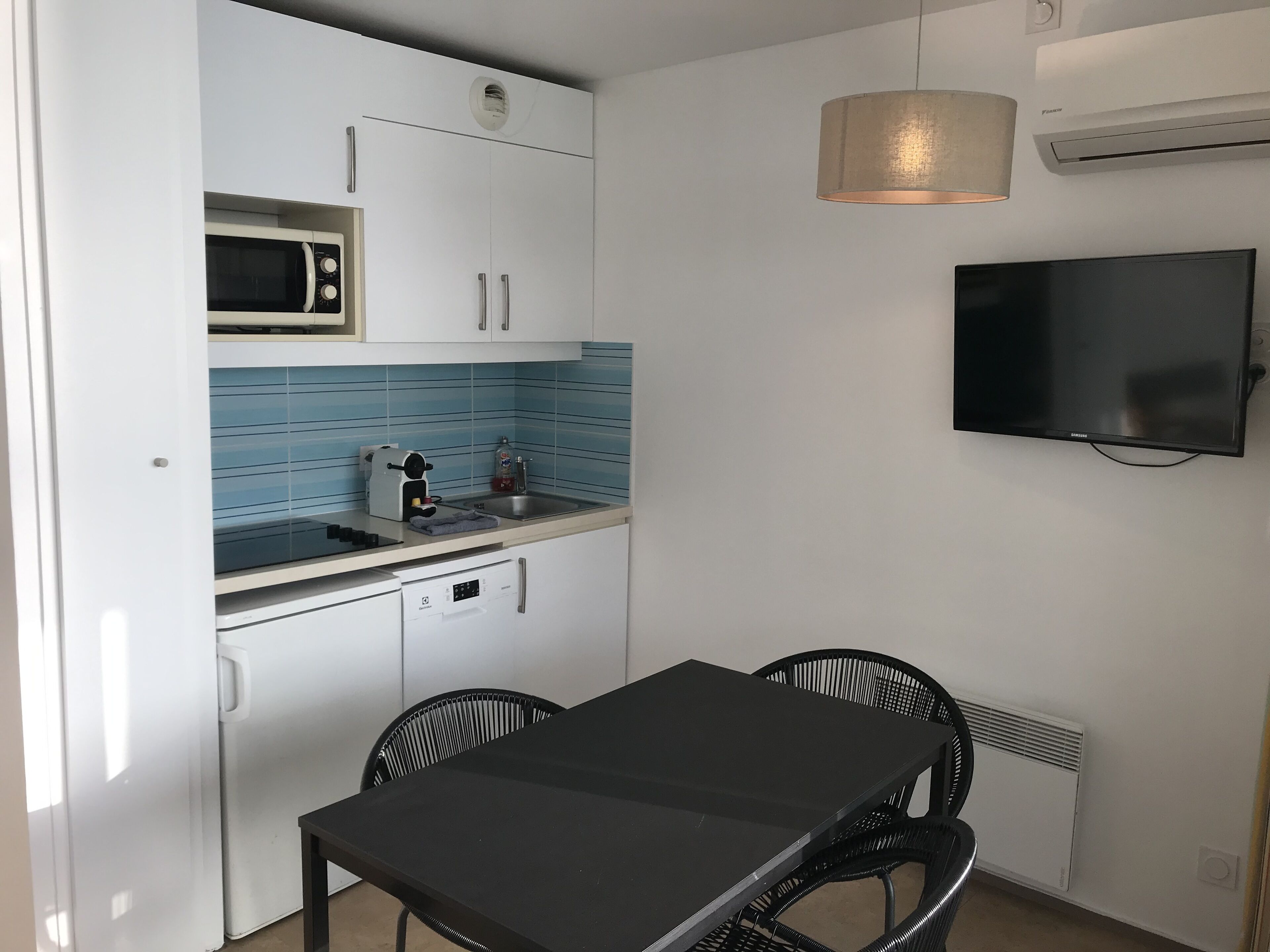 A compact kitchen with a microwave, sink, and stove, a black dining table with chairs, and a flat-screen TV mounted on the wall.