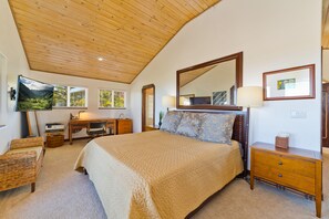 A bedroom with a large bed, wooden ceiling, a desk with a chair, a television, and a dresser.