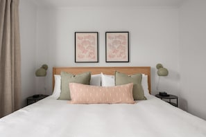 Un lit bien fait, avec une tête de lit en bois, deux tables de chevet avec des lampes et deux reproductions botaniques encadrées accrochées au mur.