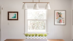 A dining area with a wooden table, white pendant lights, and a window with a view of trees. - New remodel! Walk to beach. Private heated pool & hot tub. Fenced yard. Dogs ok. (Nags Head)