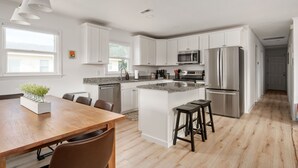 A modern kitchen with white cabinets, stainless steel appliances, a wooden dining table, and a central island with stools. - New remodel! Walk to beach. Private heated pool & hot tub. Fenced yard. Dogs ok. (Nags Head)