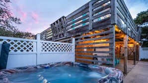 A wooden deck with a hot tub, a white fence, and a building with multiple windows. - New remodel! Walk to beach. Private heated pool & hot tub. Fenced yard. Dogs ok. (Nags Head)