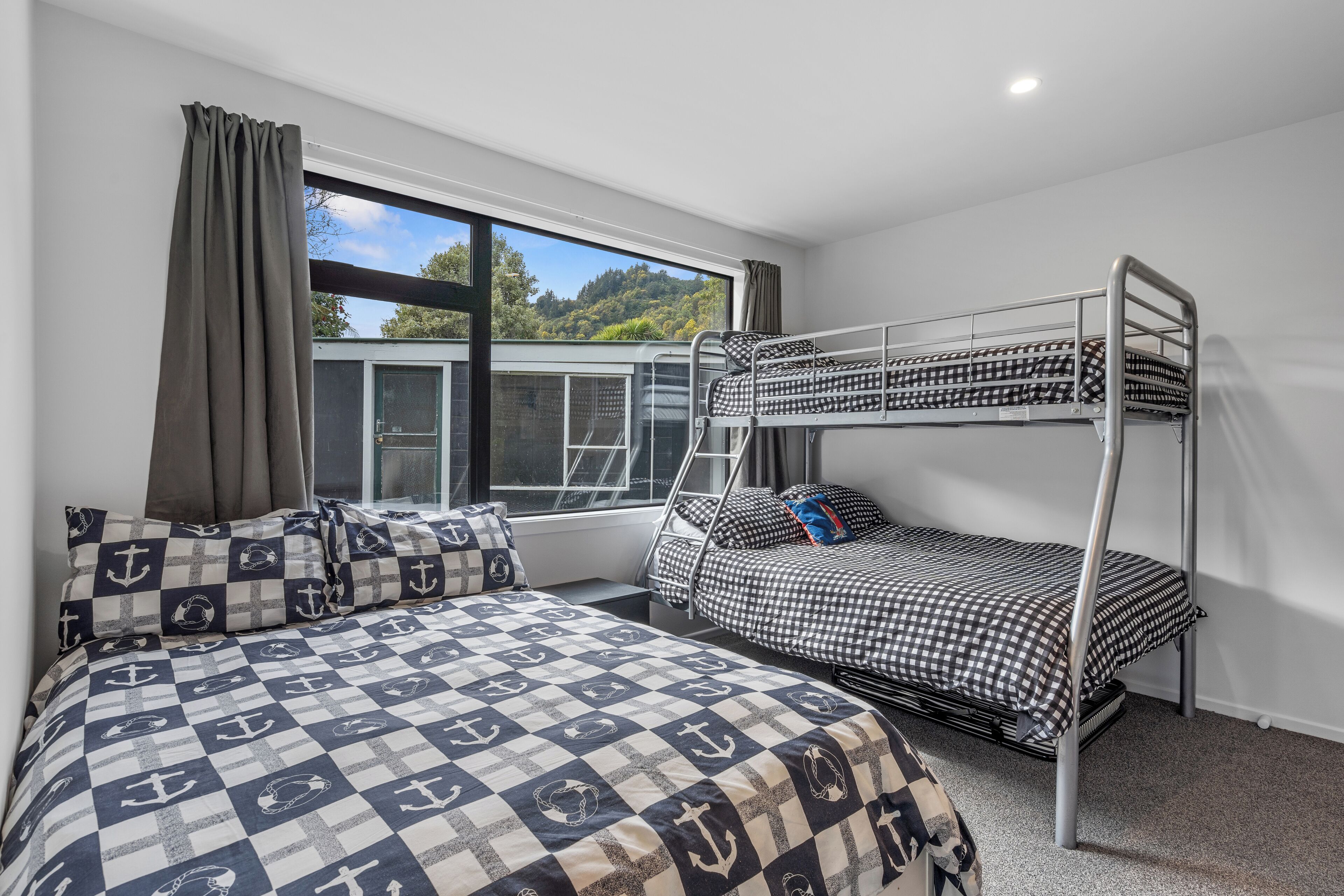 A room with a bunk bed, a single bed with patterned bedding, a window with curtains, and a view of greenery outside.