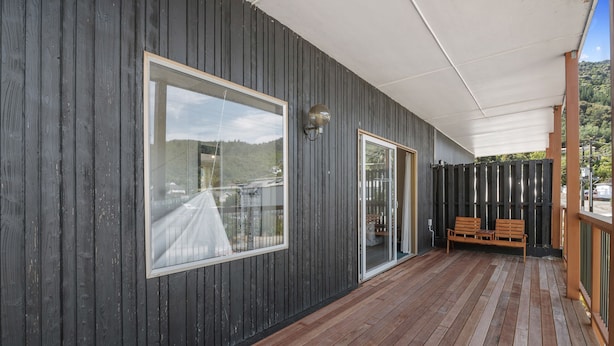 Una terraza cubierta de madera con un banco, un ventanal grande y una puerta que da a un interior.