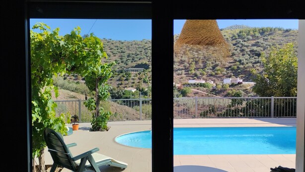 A poolside view with a clear sky, a fenced area with greenery, and a distant hilly landscape.