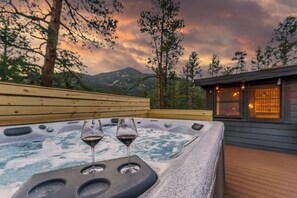 A hot tub with two glasses of red wine, surrounded by wooden deck and trees, with a mountain in the background. - Cozy Cabin w/ Hot Tub, Game Room & Private Writer's Nook - Close to town! (Evergreen)