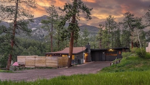 A modern cabin with a dirt driveway, surrounded by tall pine trees and a mountainous backdrop. - Cozy Cabin w/ Hot Tub, Game Room & Private Writer's Nook - Close to town! (Evergreen)