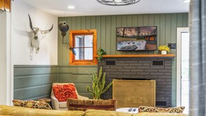 A cozy living room with a sofa, a fireplace, a mounted TV, and a wall-mounted lamp. - Cozy Cabin w/ Hot Tub, Game Room & Private Writer's Nook - Close to town! (Evergreen)