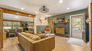 A cozy living room with a sofa, a dining area with a table and chairs, a kitchen with a refrigerator, and a fireplace. - Cozy Cabin w/ Hot Tub, Game Room & Private Writer's Nook - Close to town! (Evergreen)