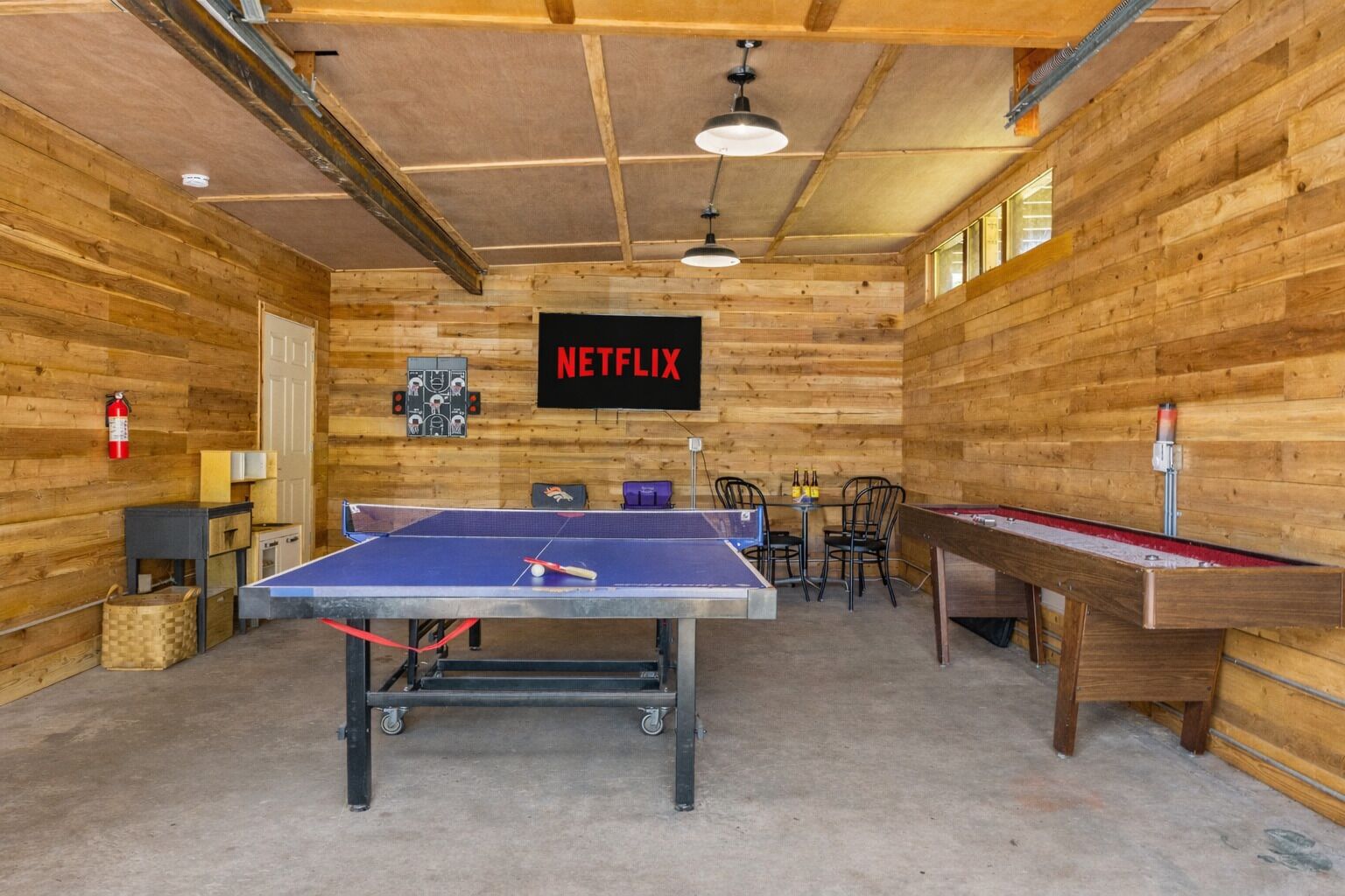 Una mesa de ping pong en una habitación con paredes de madera, una pantalla de televisión instalada mostrando 'NETFLIX' y una mesa de billar.